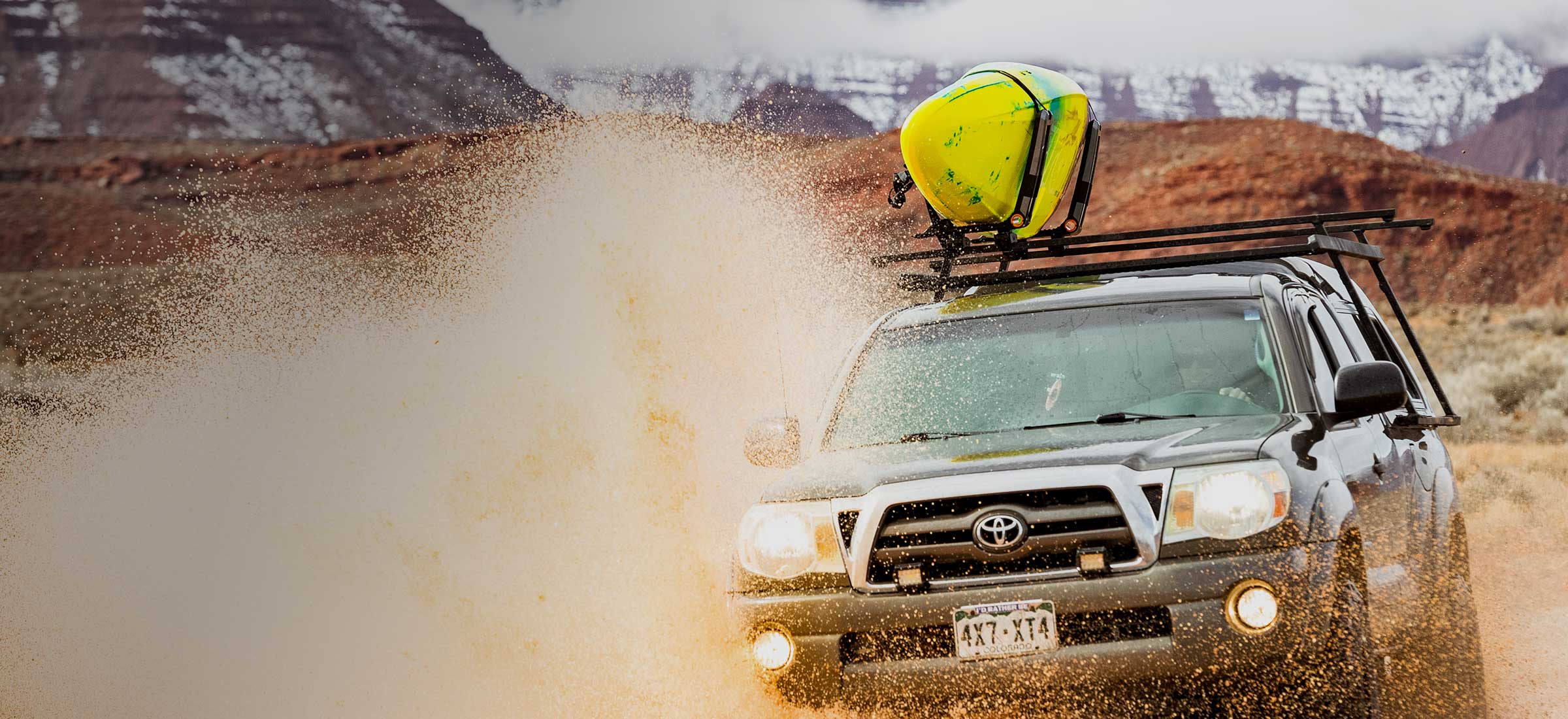 Toyota truck driving on a dirt road with a yellow kayak on the roof in a desert landscape.