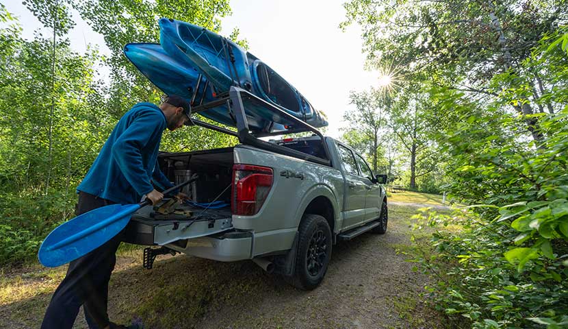 Person loading kayaks into a truck bed in a forest setting