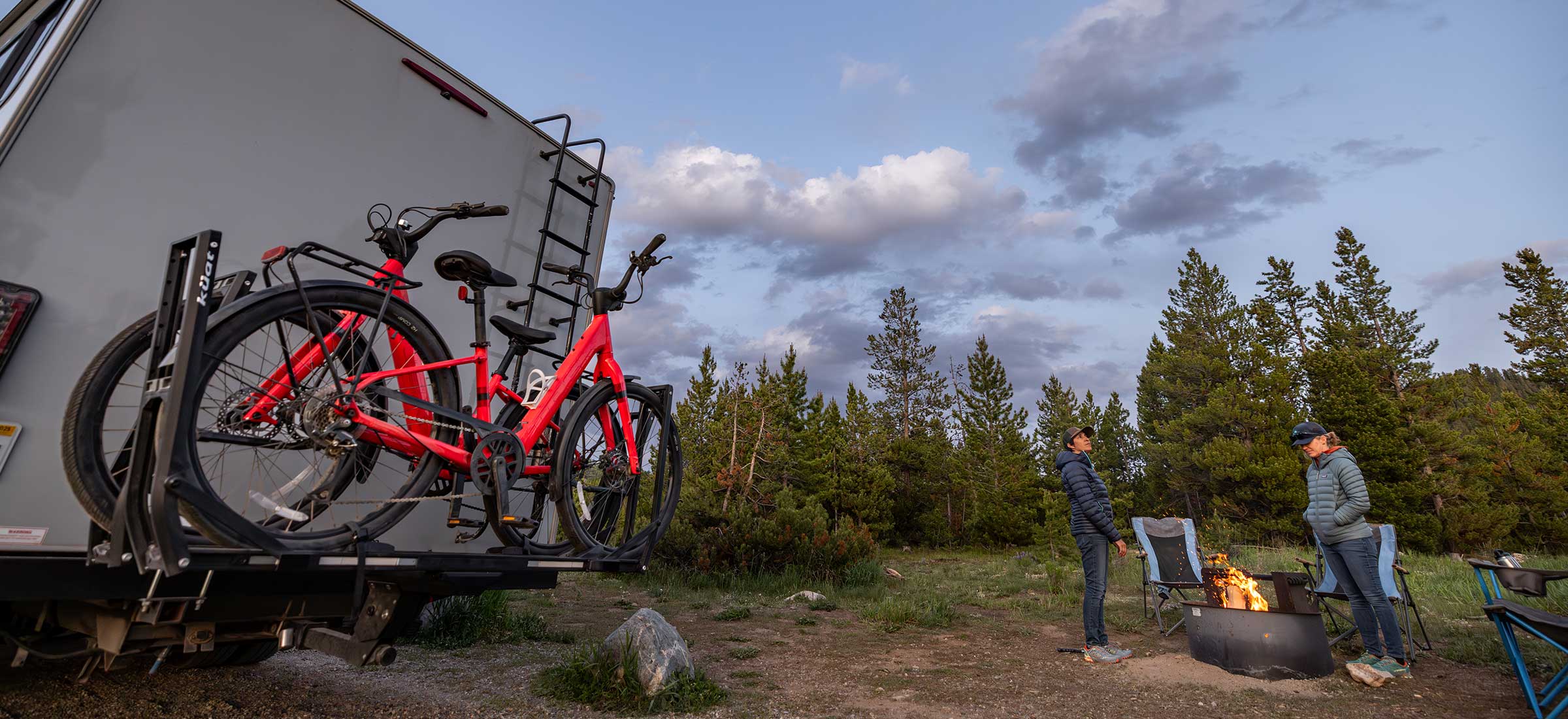 Two bicycles on a bike rack attached to an RV with two people near a campfire in a forest setting.