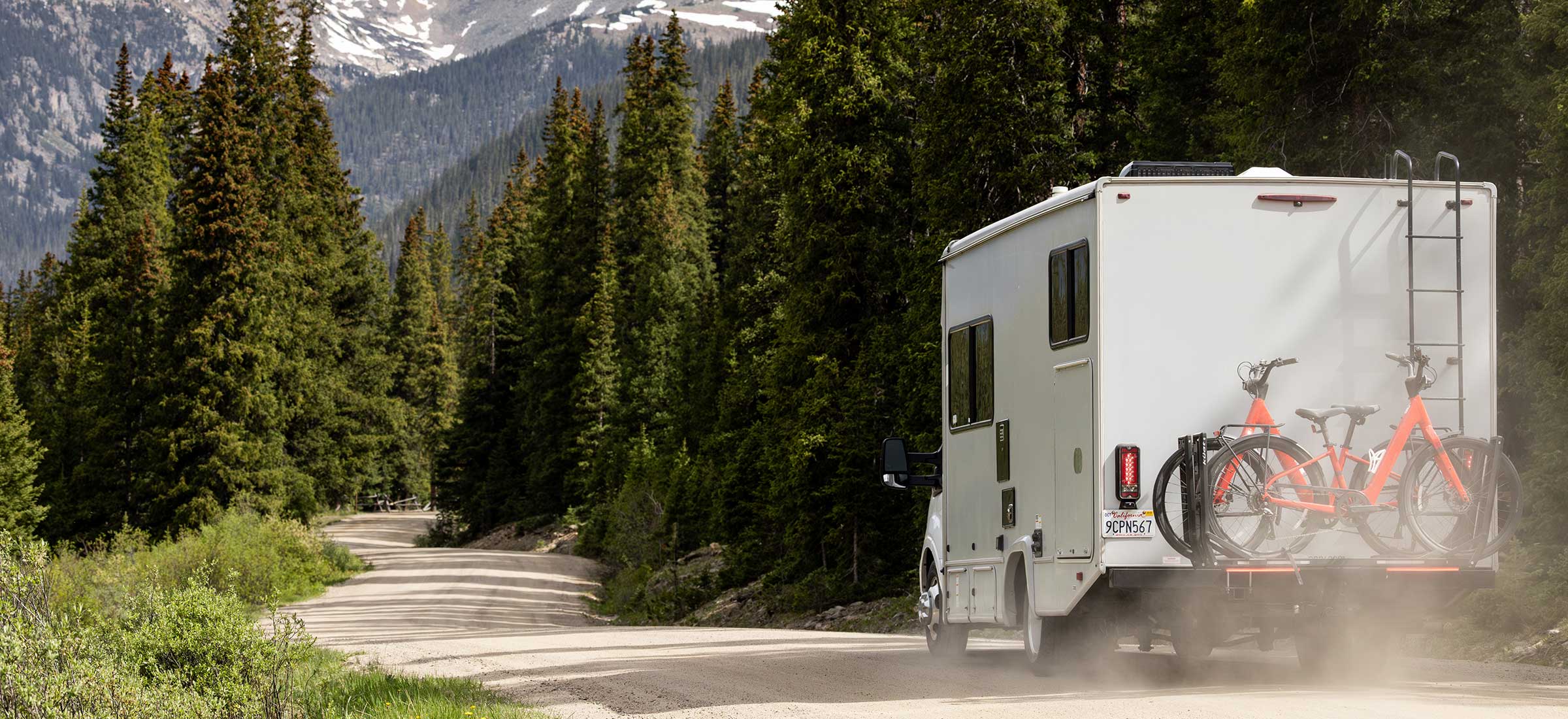RV with a bicycle attached on a road surrounded by trees and mountains