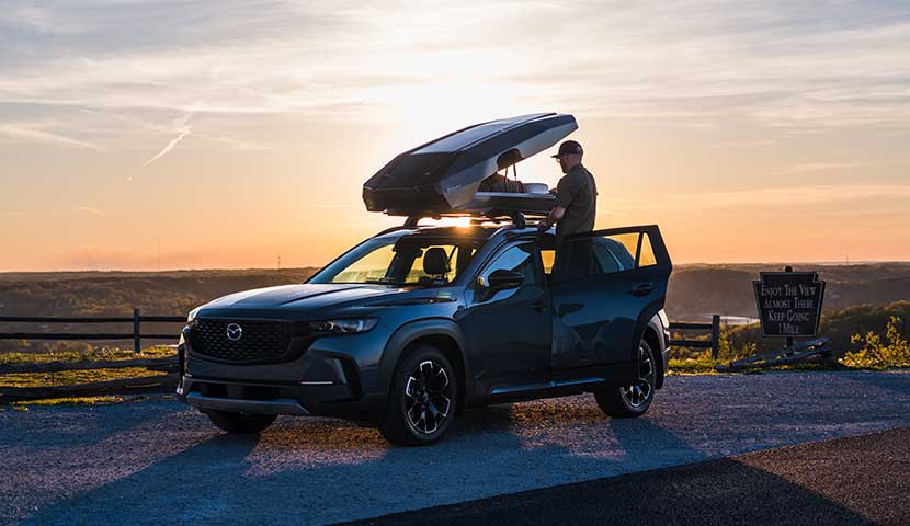 Person standing next to a Mazda SUV with an open cargo roof basket at sunset.